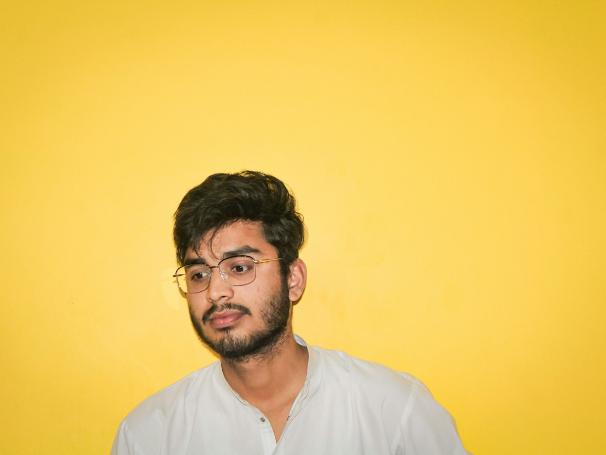 A man in round glasses against a yellow wall