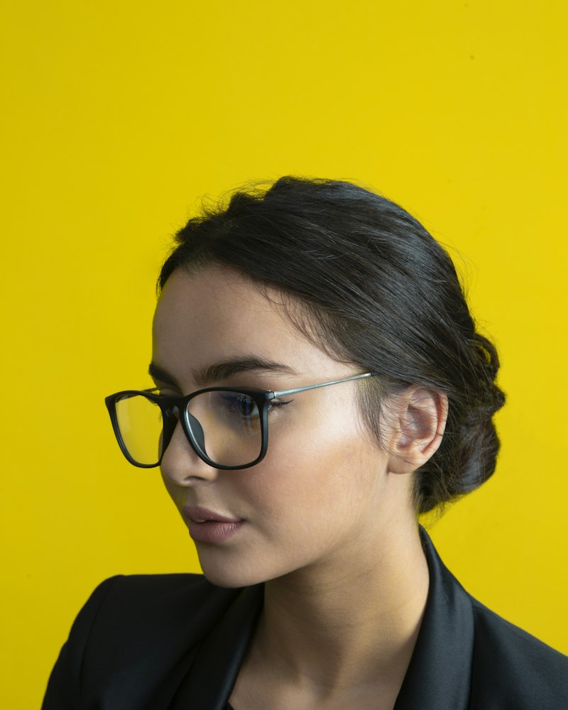 A woman in black-framed glasses against a bright yellow wall