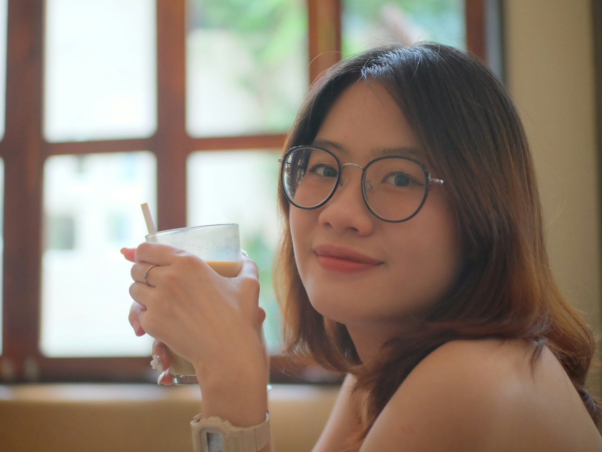 A woman in round glasses smiling by a sunny window, drink in hand